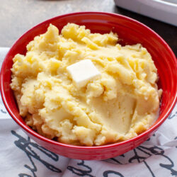 Mashed potatoes in a large red bowl with melted butter on top and other side dishes in the background