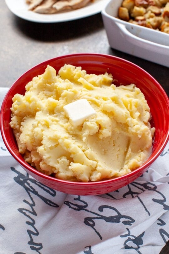 Mashed potatoes in a large red bowl with melted butter on top and other side dishes in the background