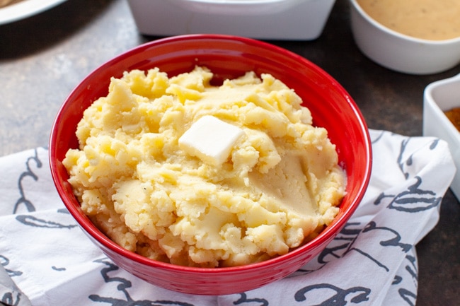 Mashed potatoes in a large red bowl with melted butter on top and other side dishes in the background