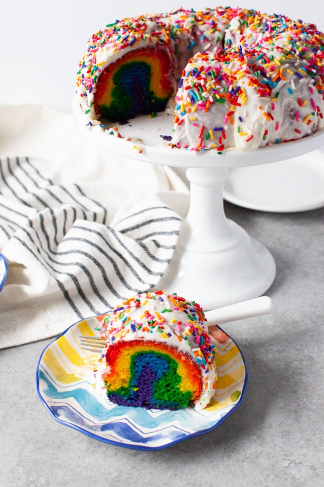 Slice of rainbow cake on a plate next to tray of cake