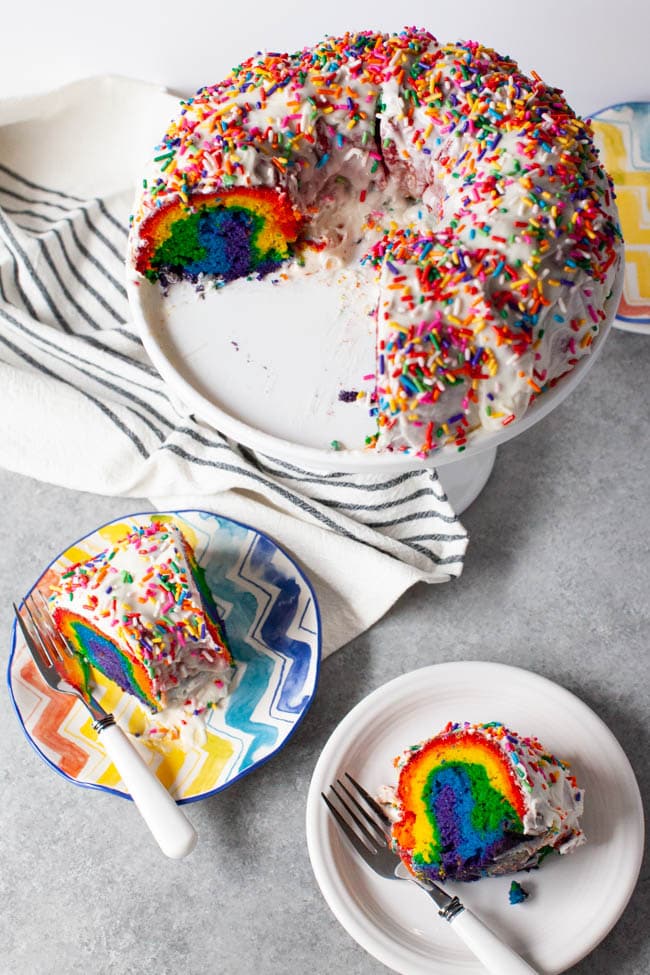 Two slices of rainbow cake on two plates with forks next to full tray of cake