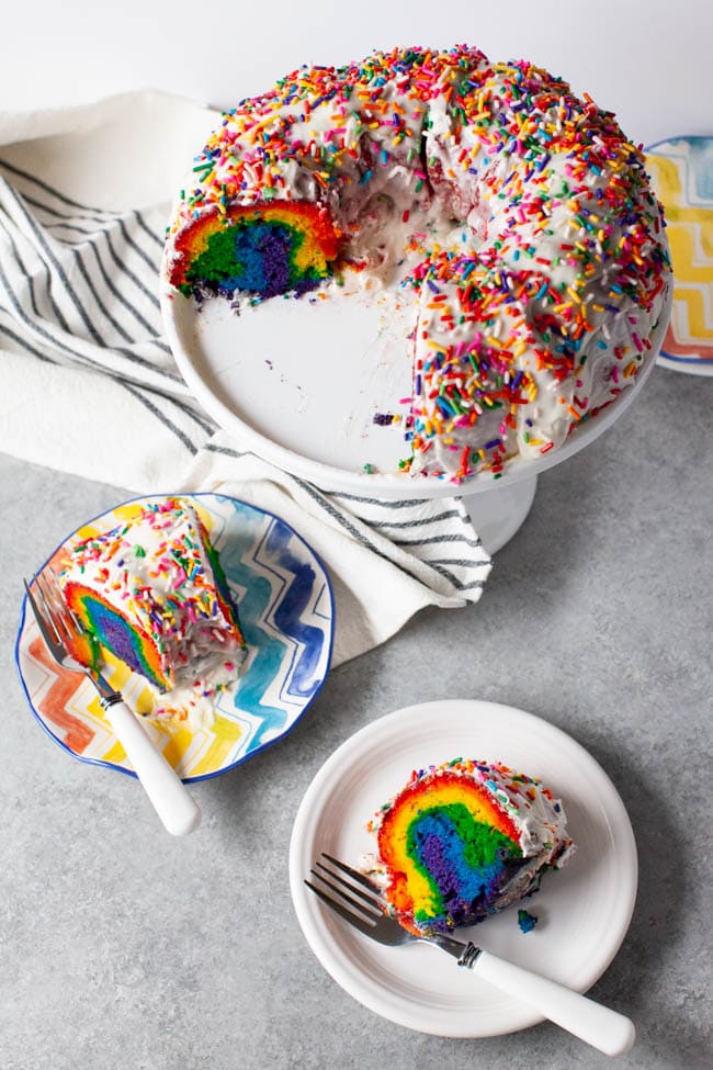 Two slices of rainbow cake on two plates with forks next to full tray of cake
