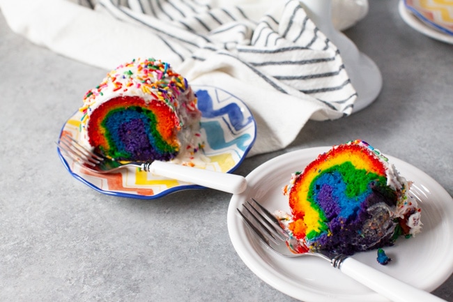Two slices of rainbow cake on two plates with forks