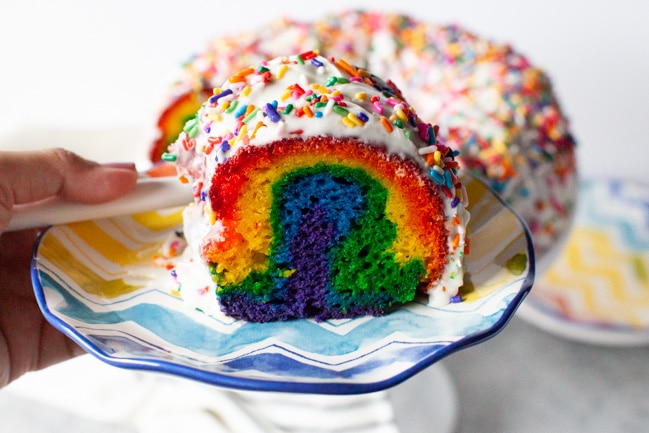 Slice of rainbow cake on a plate with the full cake in the background