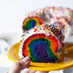 Slice of rainbow cake on a plate with the full cake in the background