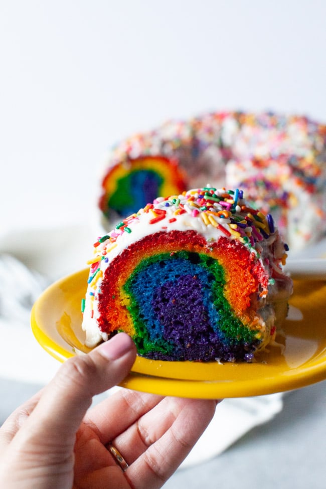 Slice of rainbow cake on a plate with the full cake in the background