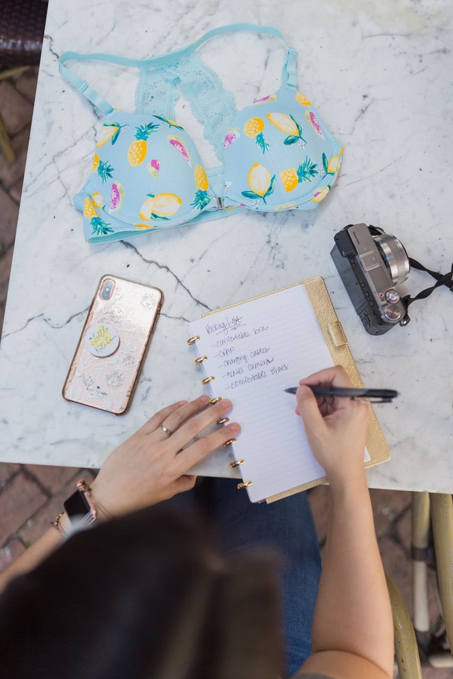 a woman writing notes on a notebook with a camera, phone and bra on a table