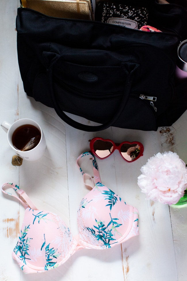 a pink bra on a table with a flower in a green vase, with sunglasses and a cup of tea and a black carry on bag