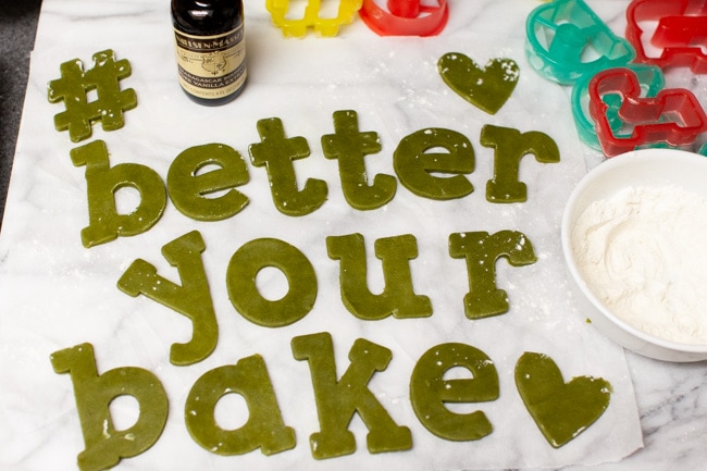 Matcha Cookie dough cut to spell #betteryourbake with bottle of vanilla, cookie cutters, and bowl of flour in background from thelittlekitchen.net