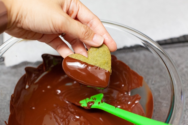 Hand dipping heart-shaped Matcha Cookies into glass bowl of melted chocolate from thelittlekitchen.net