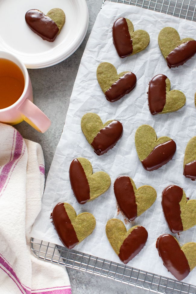 Chocolate Dipped Heart Shaped Matcha Cookies on parchment paper and cooling rack with kitchen towel, pink cup of tea, and cookie on plate in the background from thelittlekitchen.net