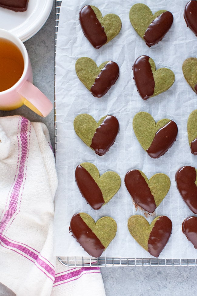 Heart Shaped Chocolate Dipped Matcha Cookies on white parchment paper, next to kitchen towel, cup of tea, and white plate from thelittlekitchen.net