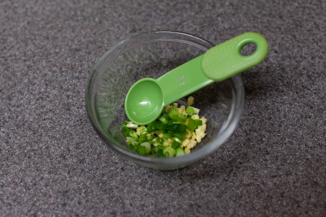 Minced ginger and green onions in a small glass bowl on a gray countertop with a green spoon