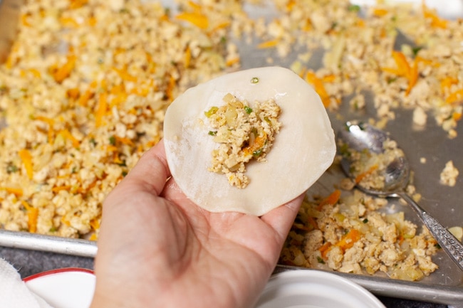 Hand holding a dumpling wrapper with filling spooned onto the middle and baking sheet of filling in background