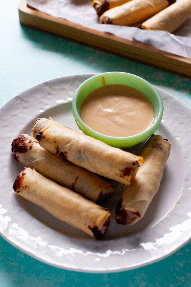 cheeseburger egg rolls on white plate with sauce, egg rolls on a tray in background