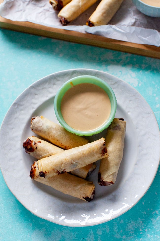 cheeseburger egg rolls on white plate with sauce, egg rolls on a tray in background