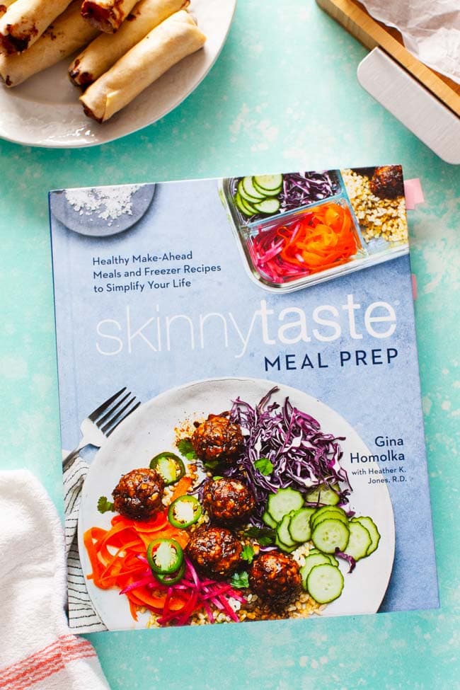 Skinnytaste Meal Prep cookbook with kitchen towel in foreground and in the background, cheeseburger egg rolls on a plate