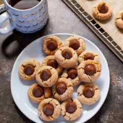 Peanut Butter Cookies topped with a Hershey Kiss sit on a off-white plate next to a cup of coffee.