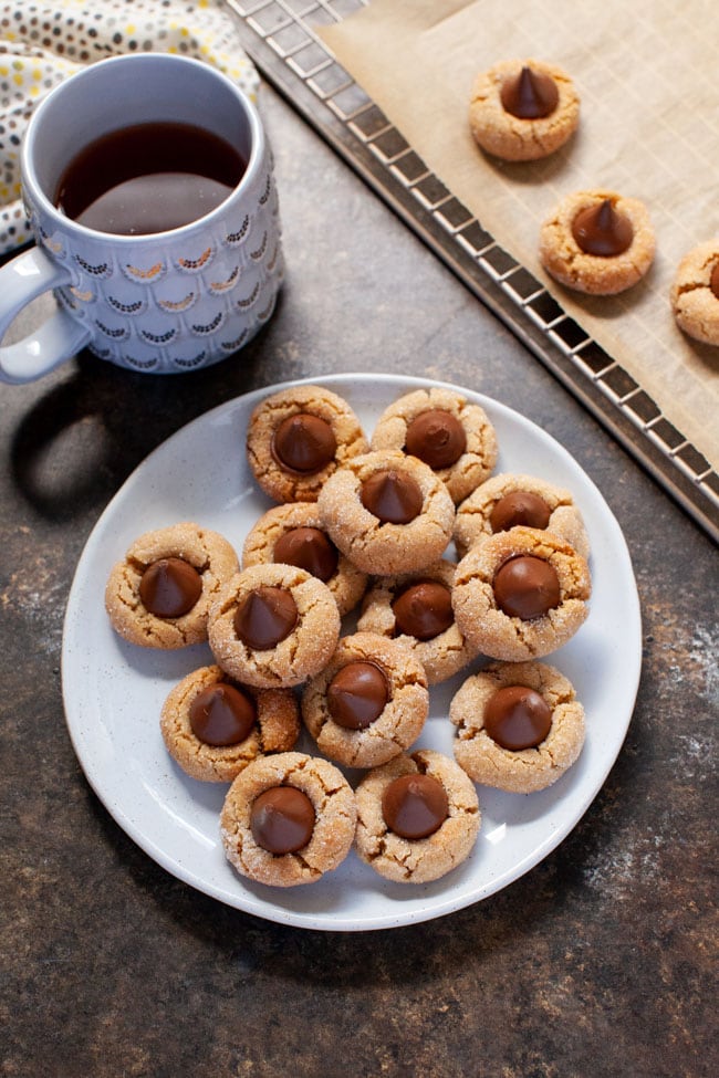 Peanut Butter Cookies topped with a Hershey Kiss sit on a off-white plate next to a cup of coffee.  
