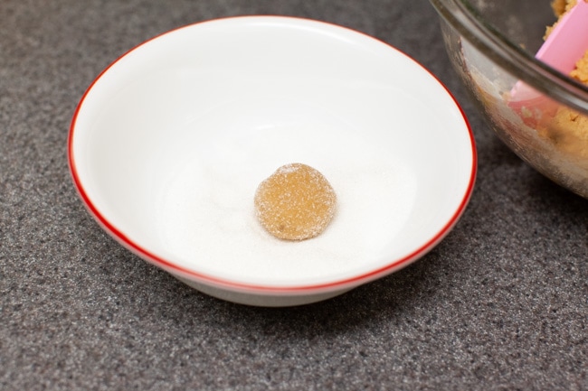 A Peanut Butter Blossom dough ball being rolled in a granulated sugar in a white bowl with red trim. A glass bowl with peanut butter blossom cookie dough can be seen in the background. 