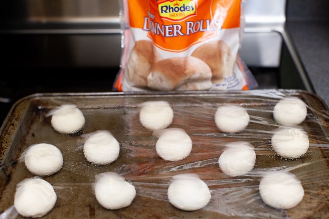 frozen dinner rolls defrosting on a baking sheet covered in plastic with a bag of Rhodes Yeast Dinner Rolls in the background