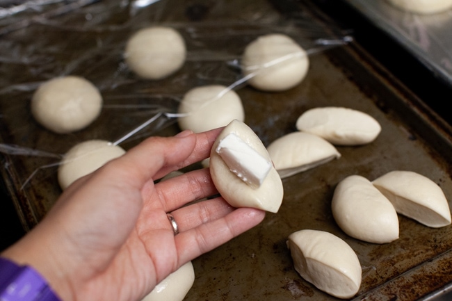 one Rhodes dinner rolls cut in half with cream cheese on a hand with other rolls on a baking sheet in the background