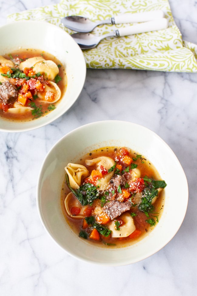 Two white bowls of Beef Tortellini Soup with two spoons and green napkin in the background.