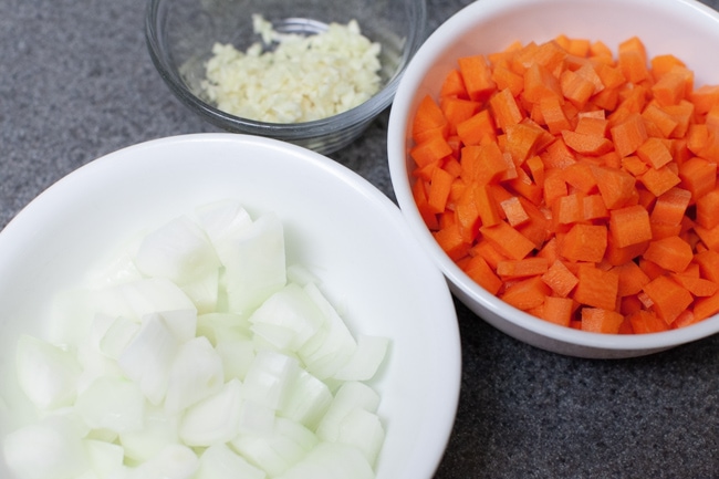 Chopped onions in a white bowl, chopped carrots in a white bowl, and minced garlic in a small glass bowl. 