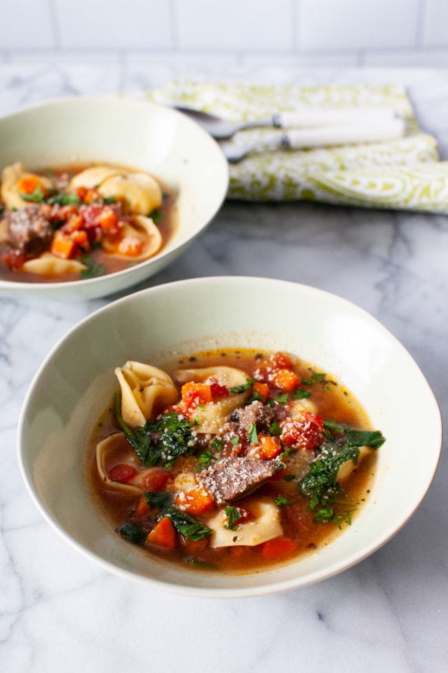 Two white bowls of Beef Tortellini Soup with two spoons and green napkin in the background.