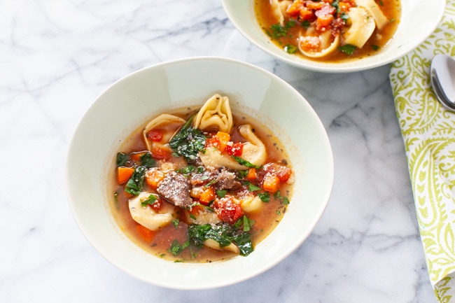 A white bowl of Beef Tortellini Soup  in the center of the picture with a green napkin and spoon in the background. 