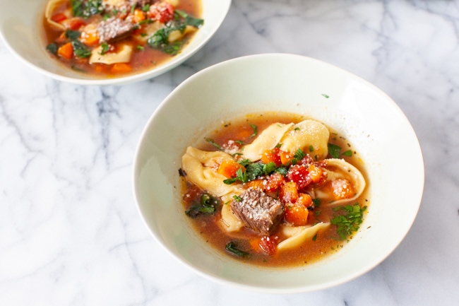 A white bowl of Beef Tortellini Soup  in the center of the picture with another bowl of soup in the background. 