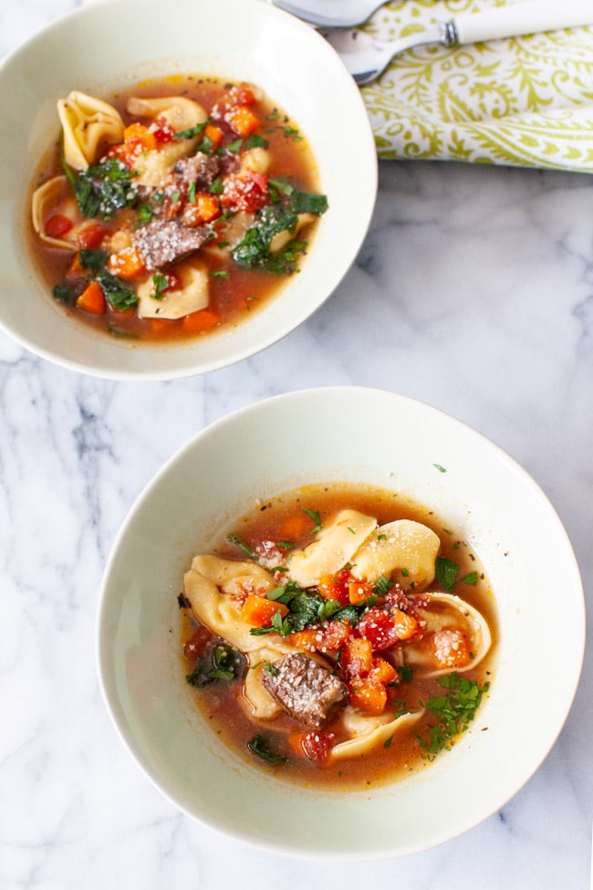 Two white bowls of Beet Tortellini Soup with a green napkin and spoons in the background. 