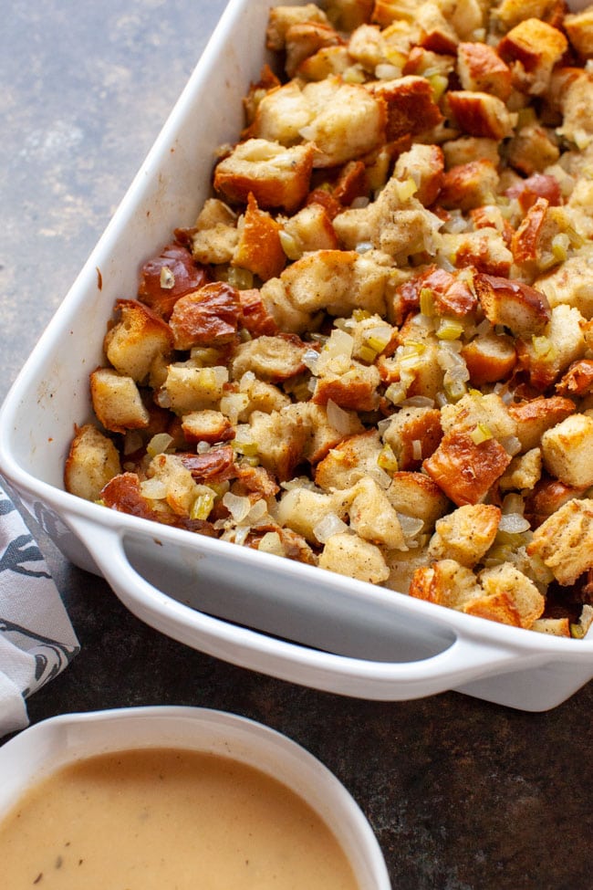 Cooked stuffing recipe in a white baking dish with a bowl of gravy in the foreground