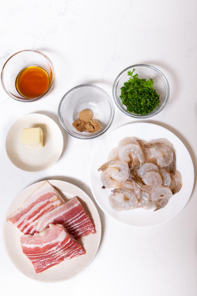 ingredients to make bacon wrapped shrimp including shrimp, bacon, butter, brown sugar, parsley, and maple syrup in individual bowls on a white background