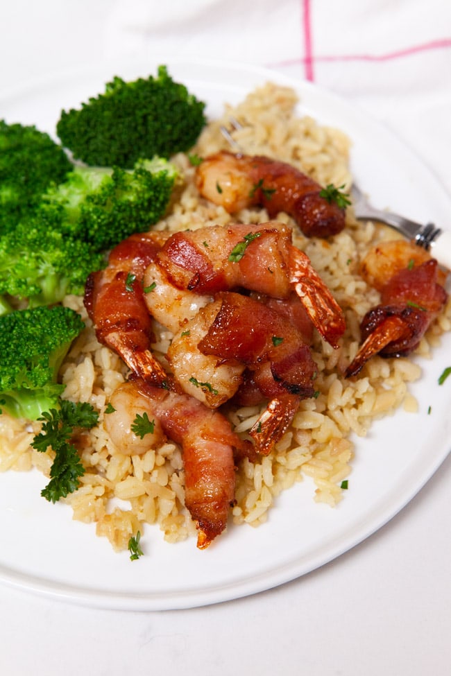shrimp wrapped in bacon con top of rice next to steamed broccoli on a white plate