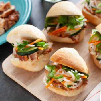 chicken banh mi sliders on a wooden board with a plate of chicken in the background