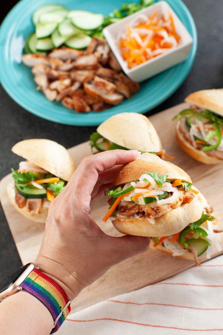 a hand holding one banh mi slider with a few other chicken banh mi sliders on a wooden board