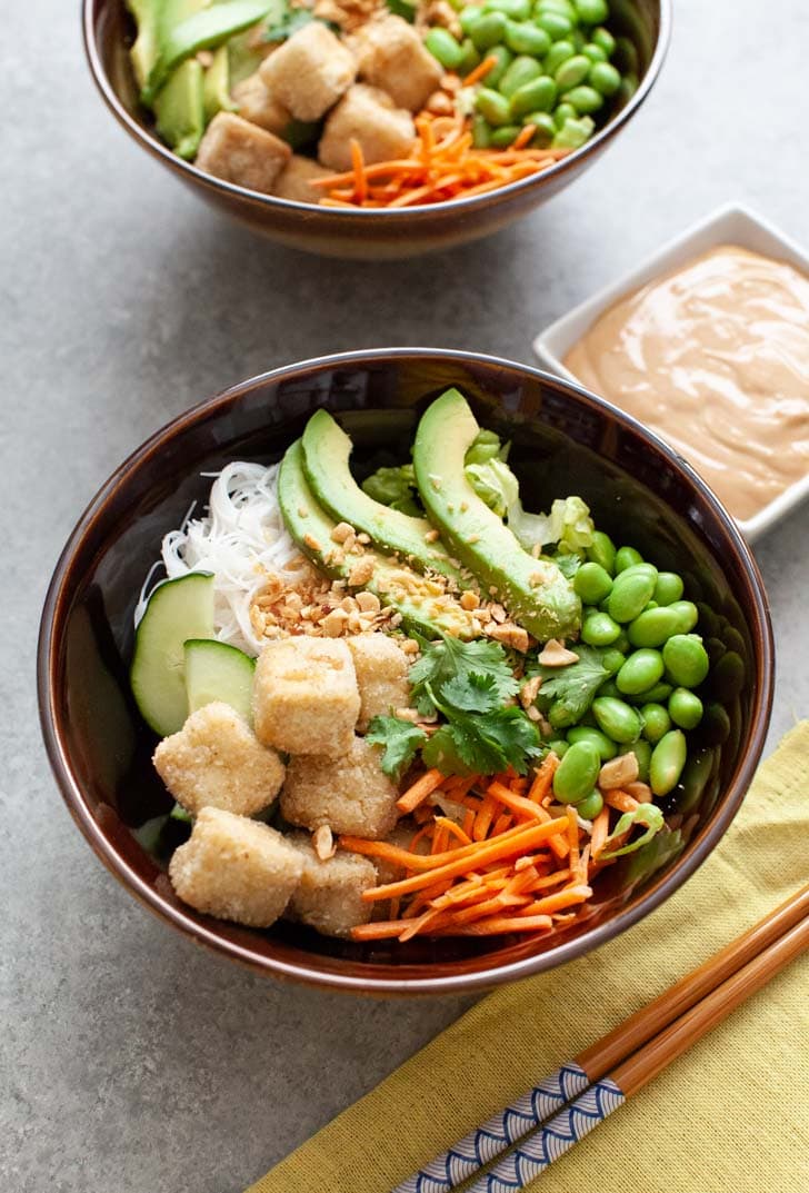 brown bowl with tofu, noodles, edamame, carrots and avocado with another bowl in the background