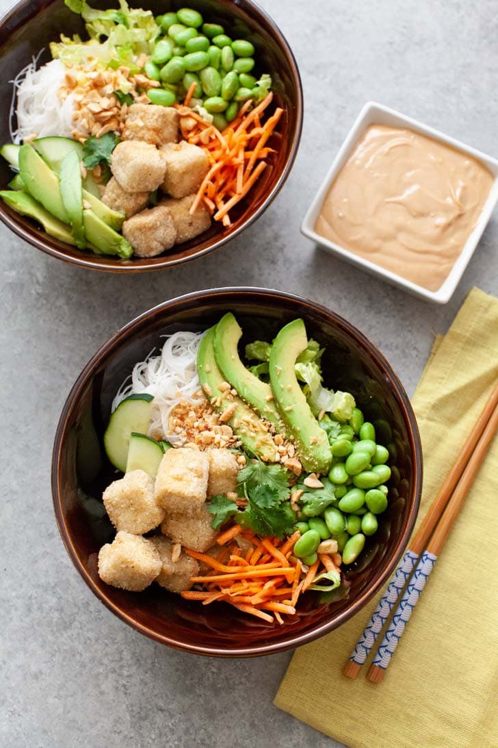 brown bowl with tofu, noodles, edamame, carrots and avocado with another bowl in the background