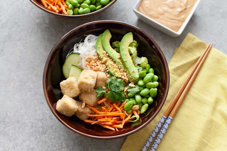 brown bowl with tofu, noodles, edamame, carrots and avocado and chopsticks on top of a napkin