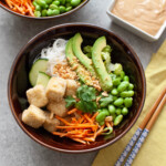 brown bowl with tofu, noodles, edamame, carrots and avocado and chopsticks on top of a napkin