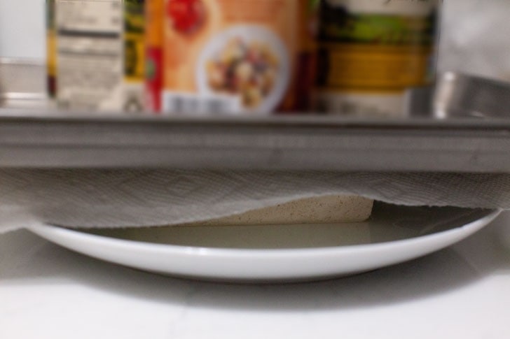 pressing tofu on a plate with a canned foods stacked on top of a cookie sheet